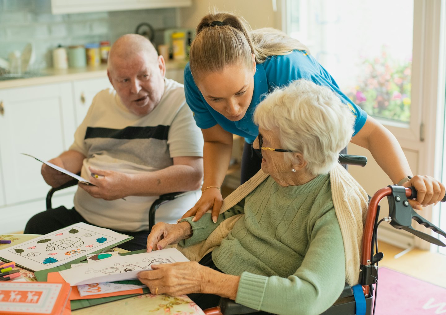 Caregiver helps elderly woman color at a table.