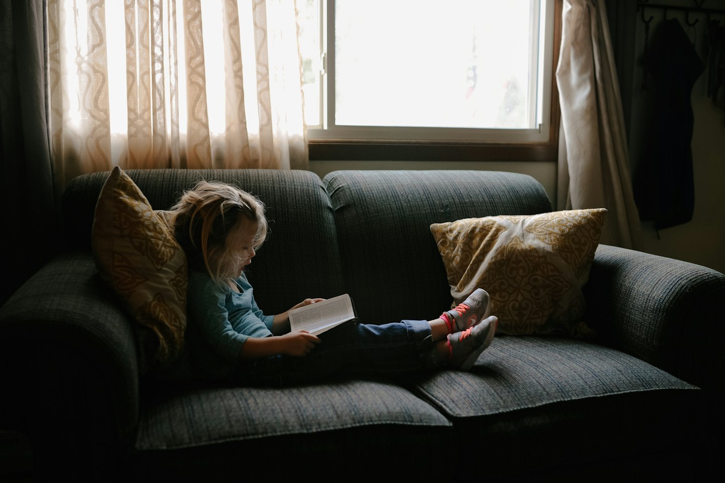 Child reads a book on a grey sofa with a yellow pillow.